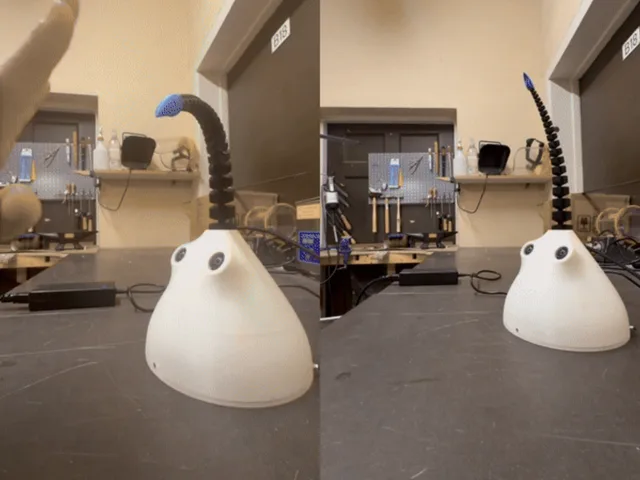The finished robot on a workbench — a white moulded dome with two eyes and a ribbed black tentacle curling up from it.
